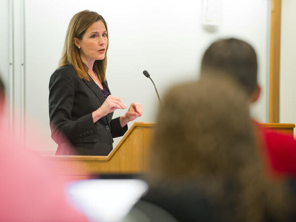 Justice Amy Coney Barrett speaks at a wooden podium in a lecture hall, gesturing with her hands. She wears a black blazer, purple top, and pearl necklace.