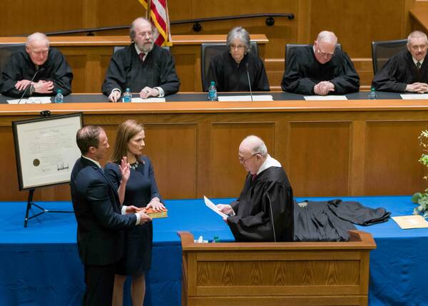 Justice Amy Coney Barrett, in a blue dress, raises her hand while her husband holds a book for her oath. Justice Clarence Thomas, in a black robe, administers the oath. Other robed justices and a US flag are visible in the wood-paneled room.