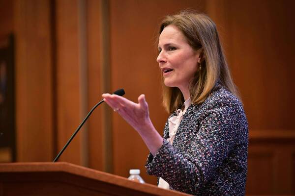 Justice Amy Coney Barrett speaks at a wooden podium, gesturing with her right hand. She wears a dark, multi-colored tweed jacket and a light blouse, her expression engaged as she talks.