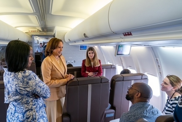 Vice President Kamala Harris, in a tan pantsuit, smiles while speaking to a woman in a blue patterned dress inside an airplane cabin. Other passengers are seen seated or standing amongst brown seats.