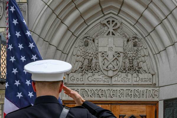A Marine in dark blue dress uniform and white cover salutes, back to viewer. A US flag hangs left. Behind are Main Building stone carvings: a shield with anchors, eagles, and "God Country Notre Dame" above wooden doors.