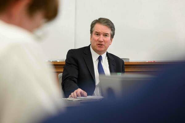 Supreme Court Justice Brett Kavanaugh speaks while seated at a table. He wears a dark suit, white shirt, and blue patterned tie, with papers in front of him.