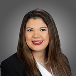 A smiling woman with long brown hair, red lipstick, a white shirt, black blazer, and pearl necklace, against a gray background.