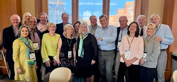 A group of 18 smiling adults, diverse in age but mostly older, pose together indoors. They wear business casual attire and name tags. The background shows large windows with a glimpse of a brick building under a blue sky.