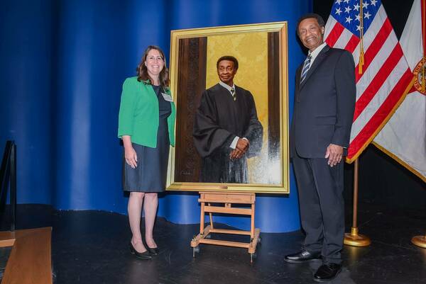 Smiling Michelle Letourneau-Belock  and Judge Charles Wilson stand beside his portrait. The painting shows Judge Wilson in a black judicial robe, with a blue and gold striped tie. An American flag and a Florida flag are in the background.