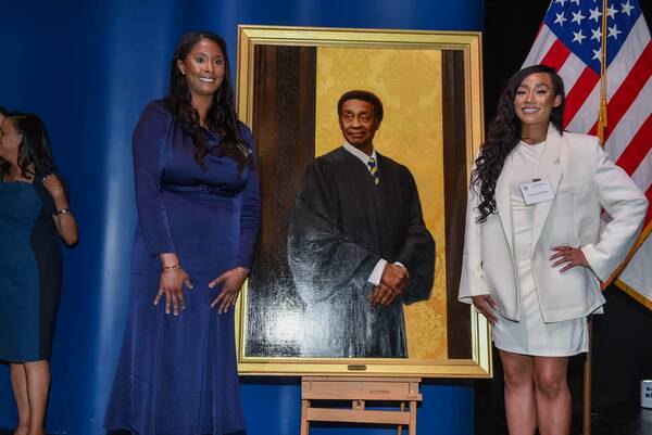 Courtney Wilson and another woman stand smiling beside a portrait of Charles R. Wilson. The portrait shows him in black judicial robes against a gold damask background, hands clasped. Courtney wears a white blazer and skirt; the other woman wears a navy dress. An American flag is behind them.