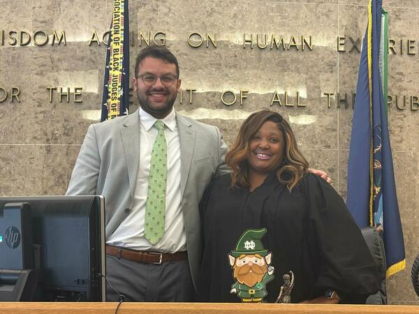 A smiling man in a gray suit and ND shamrock tie stands by a smiling woman in a black judicial robe. A Notre Dame leprechaun cutout sits before them on a desk, with a marble wall and flags in the background.