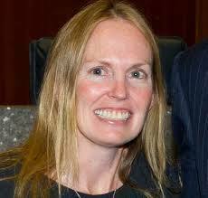 A blonde woman with a wide, bright smile looks forward, wearing a dark shirt.