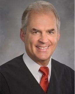 A smiling man with gray hair in a black judicial or academic robe and a red tie.
