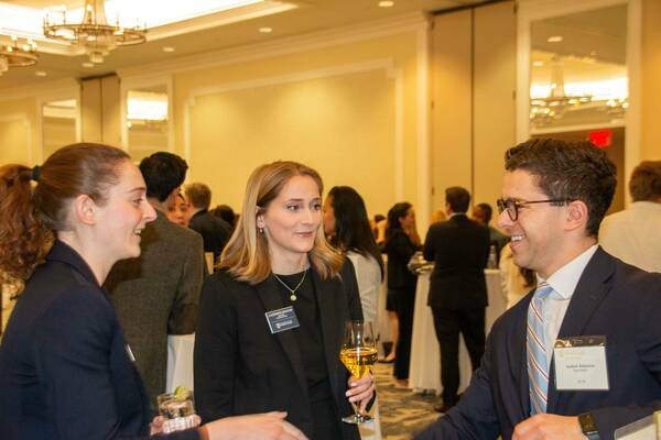 Andrew Athenson (right, navy suit, glasses) smiles at Catherine Kolesar (center, black blazer, holding wine), who smiles back while conversing with another woman (left, dark blazer, holding a drink). All three smile. Name tags show Athenson is JD '24, and Kolesar is with Notre Dame.