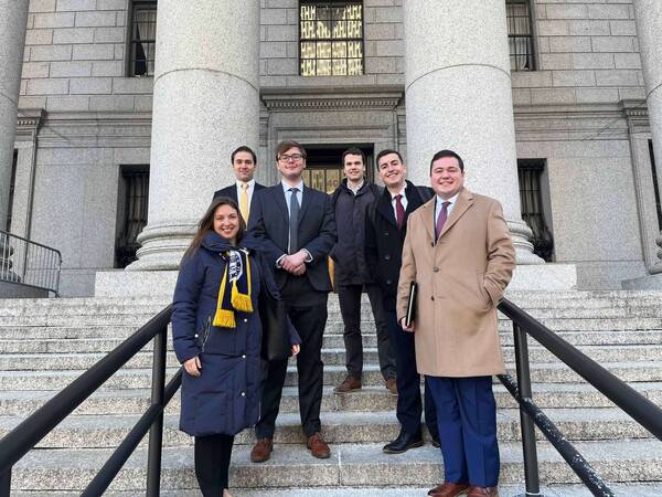 Six smiling individuals, five men and one woman, stand on the granite steps of a grand stone building with classical columns. The woman wears a blue coat and blue-and-gold Notre Dame scarf. The men wear suits and coats, some with ties, suggesting a professional or academic outing.