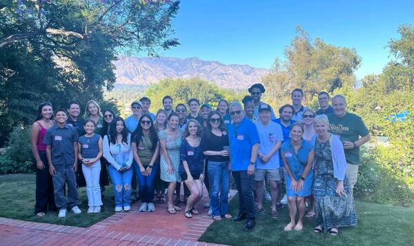 A diverse group of smiling people of varying ages stands on a green lawn next to a brick path. Lush trees and distant mountains are in the background under a clear blue sky. Many wear casual attire and name tags.