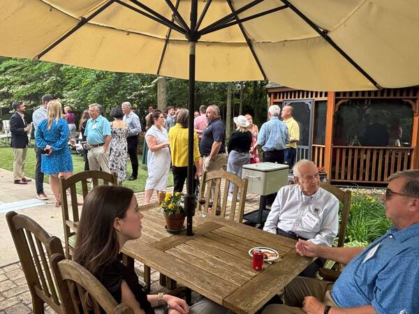 An outdoor gathering under a yellow umbrella. In the foreground, a young woman and older man with glasses sit at a wooden table with a red soda can and yellow flowers. Guests, including a woman in a blue polka-dot dress, mingle on a lawn and paved patio in the background.