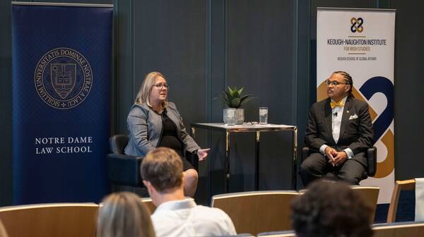 A blonde woman in glasses and a black-and-white plaid jacket speaks, gesturing. Beside her, a man with dreadlocks and glasses, wearing a dark pinstripe suit and yellow bow tie, listens. Banners for Notre Dame Law School and the Keough-Naughton Institute are behind them.