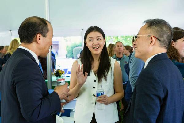 A smiling woman in a white sleeveless vest holds a water bottle and gestures while conversing with two men in dark suits. The man on the left holds a silver can; the man on the right wears glasses and smiles. They are under a white tent at an outdoor event.