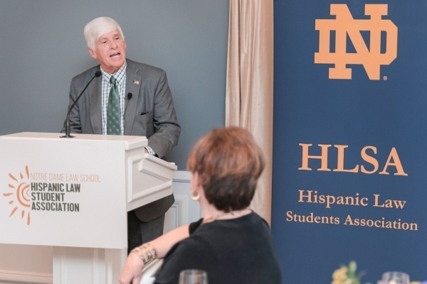 A man with white hair, in a gray suit, green tie, and US flag lapel pin, speaks from a podium displaying the Notre Dame Law School Hispanic Law Student Association logo. A blue banner with the gold ND logo and "HLSA" is behind him.