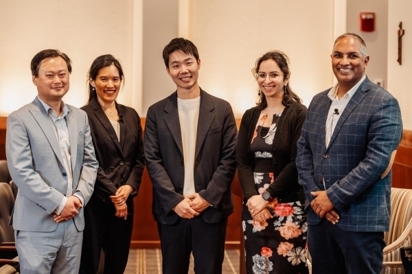 Five diverse, smiling professionals – three men and two women – stand in business attire, two with lapel mics. One man wears a light blue suit, another a blue plaid jacket. A woman wears a black floral dress. Background is light walls with wood paneling.