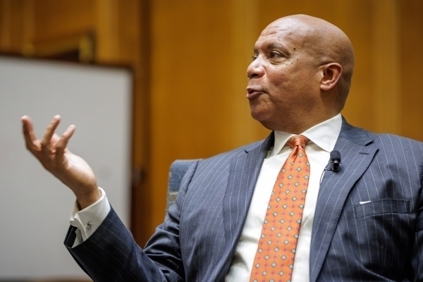 Bald man in a dark pinstripe suit, white shirt, and orange patterned tie speaking, gesturing with his left hand raised. A lapel microphone is clipped to his suit jacket, with a warm wood-paneled background.