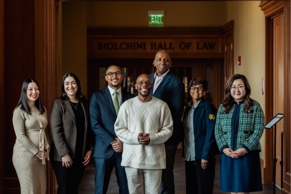 Six smiling people, three men and three women, pose in Biolchini Hall of Law. They wear varied business casual attire, including suits, sweaters, and a jacket with a Notre Dame logo.