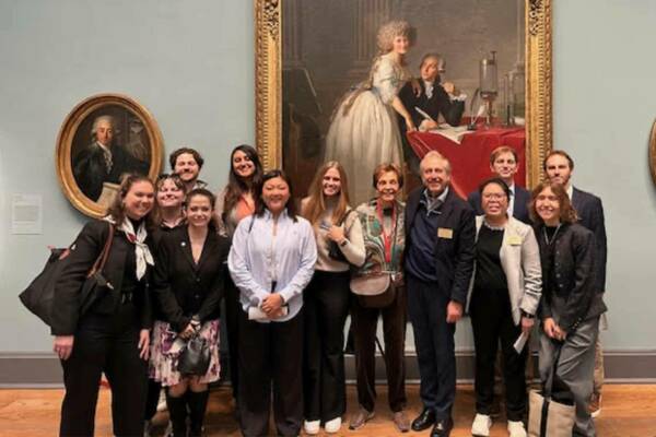 A diverse group of smiling men and women pose in an art museum, standing before a large classical portrait of a woman with an elaborate hairstyle and a man writing at a red table.