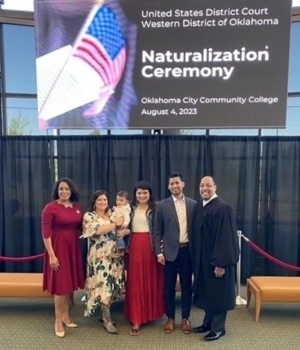 A smiling group, including a judge in a black robe, poses before a screen showing an American flag and "Naturalization Ceremony, Oklahoma City Community College, Aug 4, 2023." A woman in a red dress, a woman holding a baby in a floral dress, a woman in a red skirt, and a man in a suit are in the group.