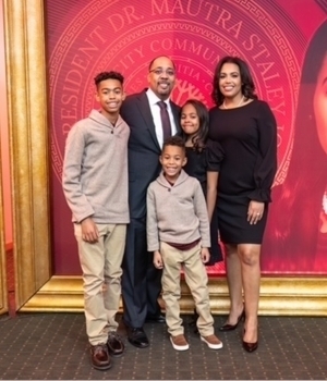 A family of five smiles: a man in a dark suit, a woman in a black dress, two boys in tan long-sleeve shirts, and a girl in a dark dress. They stand together in front of a red event banner with gold lettering.