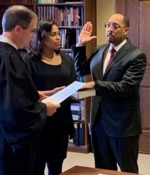 A man in a dark suit with glasses raises his right hand to take an oath, his left hand on a white book. A man in black judicial robes holds a document, while a woman in a black dress and pearls observes. A bookshelf is in the background.