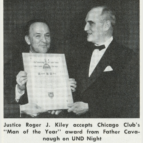 Justice Roger J. Kiley, smiling in a dark suit, holds a certificate. Father Cavanaugh, in a dark suit and bow tie, smiles as he presents the award.