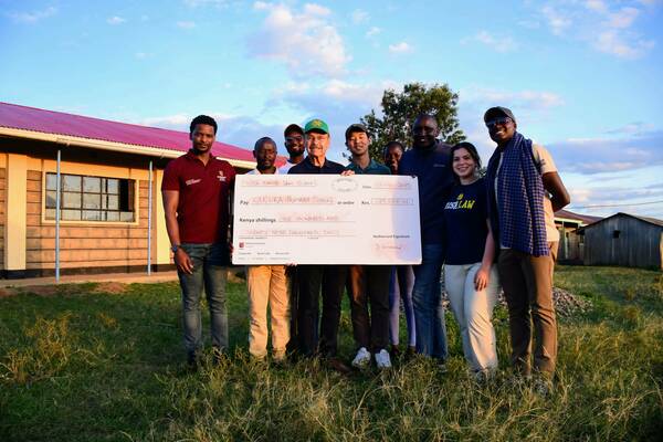 Eight smiling people, including a man in a green Notre Dame hat and a woman in an "ND Irish Law" shirt, hold a large check outdoors. The check is from Notre Dame Law School to Karuka Primary School for 129,000 Kenya shillings. Simple buildings under a cloudy sky.