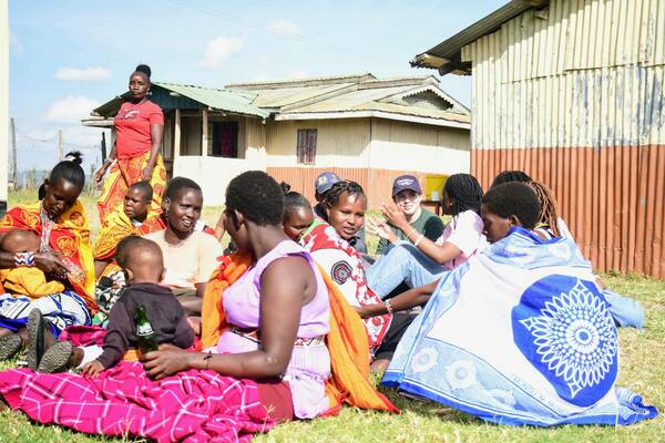 A diverse group of women and children sit on grass outdoors. Many wear vibrant red, yellow, and blue patterned clothing. One woman feeds a baby. A student in a baseball cap smiles while conversing with a local woman, gesturing with her hands. Corrugated metal buildings are in the background.