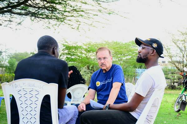 Three men sit in white chairs outdoors. An older man with glasses and a mustache wears a blue Notre Dame shirt, looking attentive. A younger man in a white shirt and black cap listens. Another man faces away, also listening.
