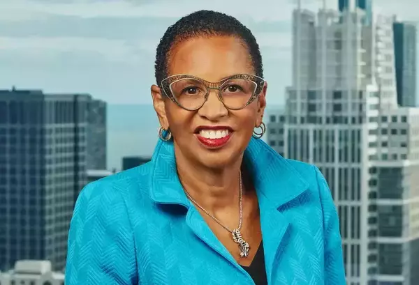 Smiling Black woman with short curly hair, wearing a turquoise blazer and ornate cat-eye glasses, cityscape background.