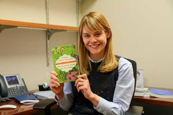 A blonde woman smiles, holding up a green copy of Gabriel García Márquez's "One Hundred Years of Solitude."