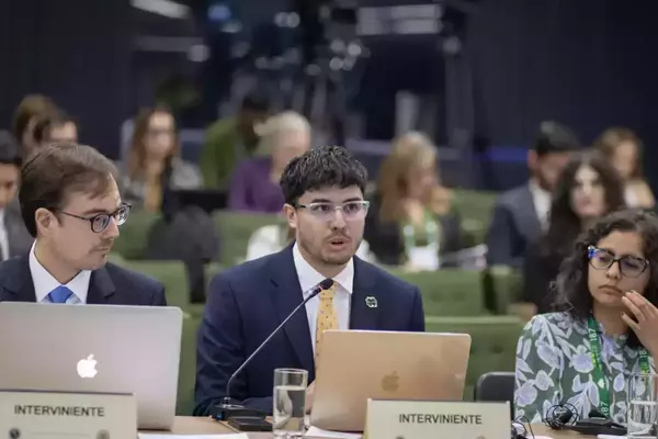 Young man in navy suit, yellow tie, and clear glasses speaks into a microphone at a conference table with an "INTERVINIENTE" sign.