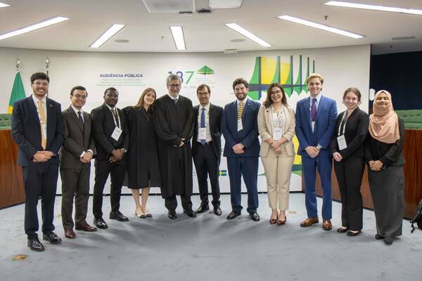 Eleven people, including one in a black judicial robe, smile in a formal room with "Public Hearing" on the wall and a Brazilian flag.