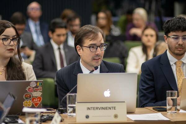 A man in a dark pinstripe suit and glasses speaks into a microphone at a table with an 'INTERVINIENTE' name card.