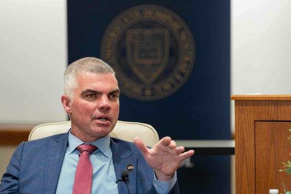 Gray-haired man in blue plaid suit, red tie, speaks with open mouth, gesturing left hand. Blurred Notre Dame seal in background.