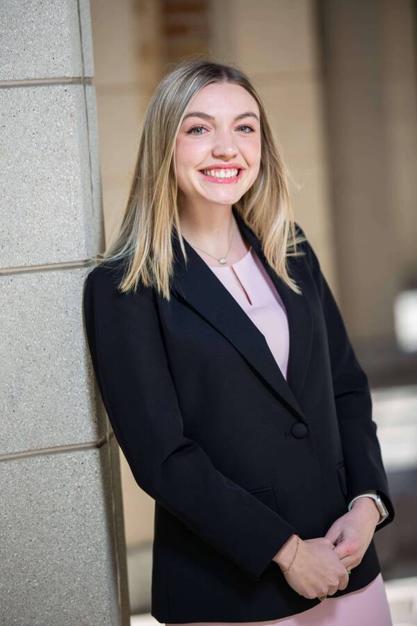 Nikki Rafferty, a young woman with blonde hair and green eyes smiles brightly, wearing a black blazer and pink top, hands clasped, leaning on a grey column.