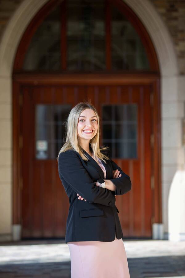 Nikki Rafferty, a smiling blonde woman in black blazer and pink skirt, arms crossed, stands before an arched dark wood door at Notre Dame.