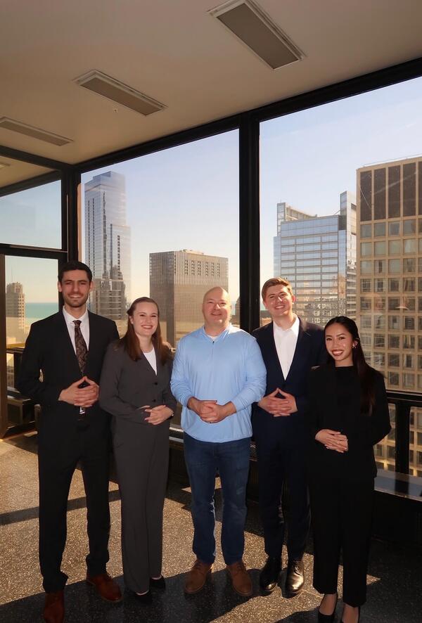Three men and two women, smiling, stand in business and casual clothes against a city skyline with Lake Michigan.