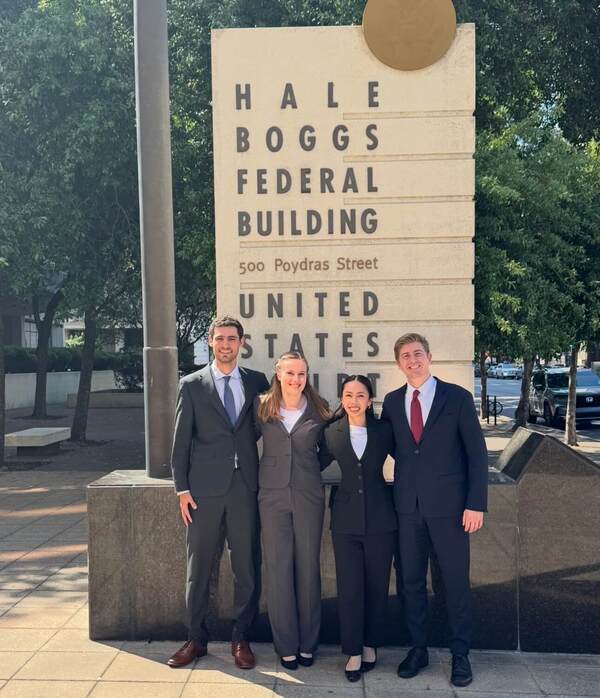 Four smiling young adults in suits pose before the Hale Boggs Federal Building sign. Two men in dark suits with ties, two women in suits.