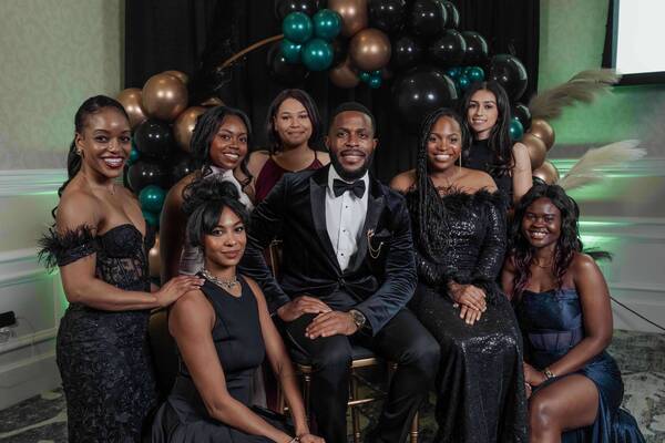 Eight smiling people, a man in a tuxedo and seven women in formal dresses, pose before a black, teal, gold balloon arch.