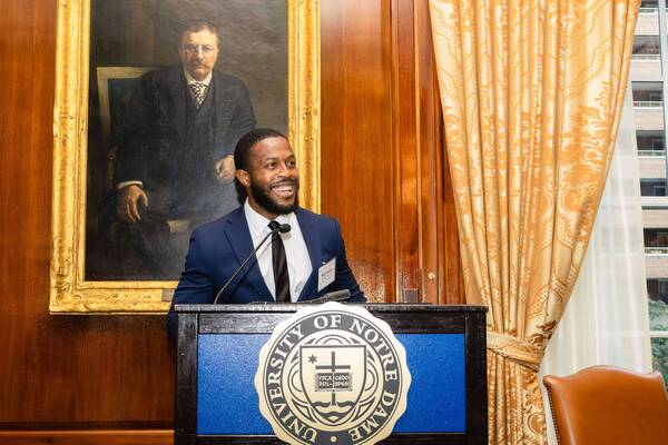 Smiling man in blue suit speaks at a podium with the Notre Dame seal. A portrait of Theodore Roosevelt hangs behind him.