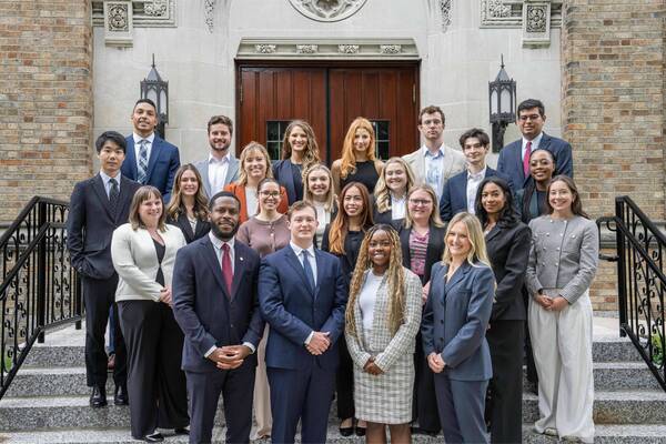 Diverse group of young adults in professional attire smiles on stone steps of an ornate Notre Dame building.