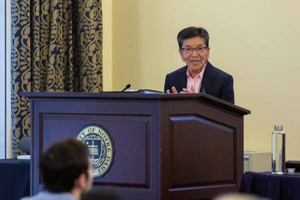 Smiling woman in glasses, dark suit, pink shirt gestures while speaking at a University of Notre Dame podium.