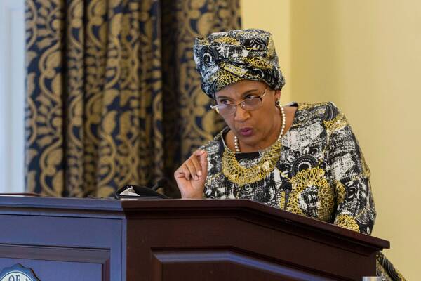 A woman in a black, white, gold patterned headwrap and dress speaks from a dark podium, gesturing with her right hand.