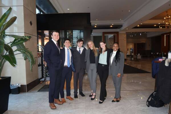 Six smiling young adults in business attire. Three men wear dark suits; three women wear grey blazers, black pants, and blouses.
