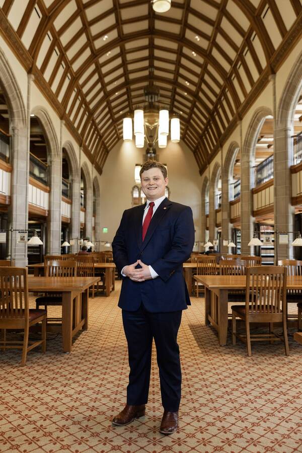 Smiling man in navy suit, red tie, brown boots, hands clasped, stands in Hesburgh Library's grand reading room.