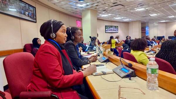Two women in headphones sit focused at a conference table. One wears a red jacket and black head covering, writing.