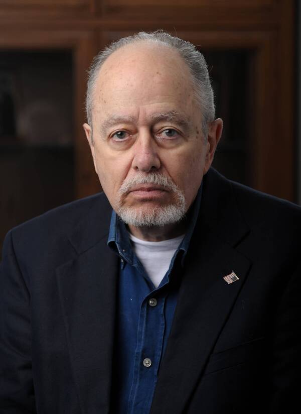Older man with gray hair, beard, and mustache, wearing a dark blue blazer, blue shirt, and US flag lapel pin.